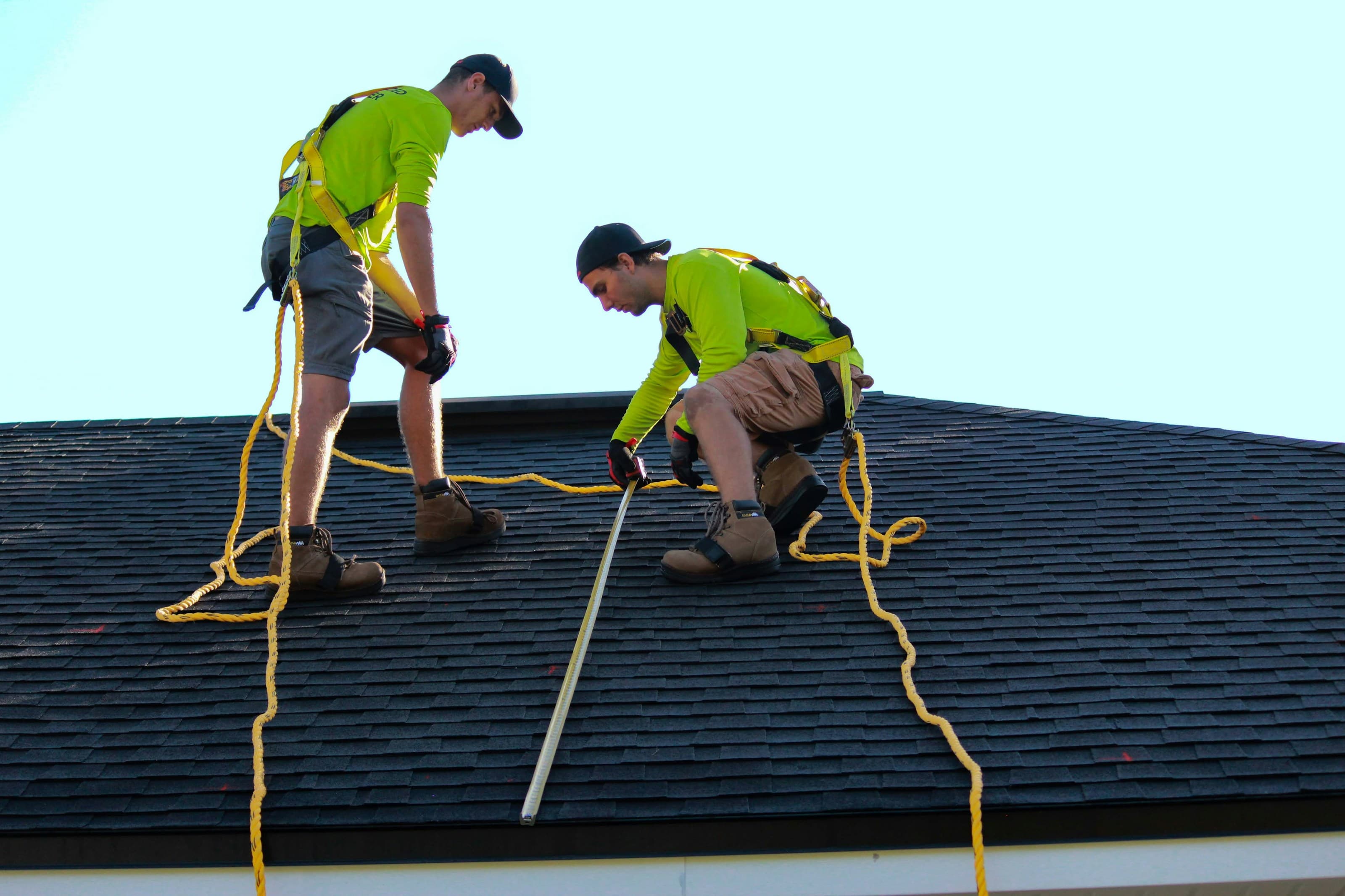 Two roofers in safety harnesses measuring along a dark shingle roof